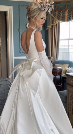 a woman in a white wedding dress is standing near a window with a chandelier