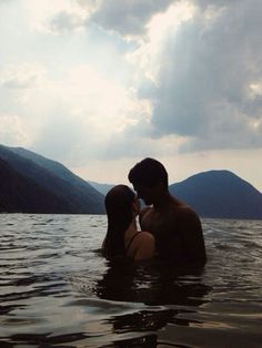 two people are in the water with mountains in the backgroung and clouds in the sky