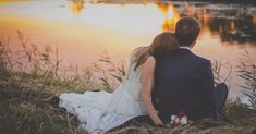 a bride and groom are sitting on the grass by the water at sunset or sunrise