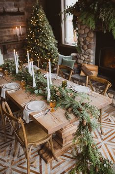 a wooden table topped with white plates and greenery next to a fireplace filled with lit candles