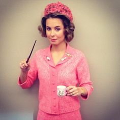 a woman in pink is holding a coffee cup and a black stick while standing against a wall