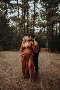 a pregnant couple standing in the middle of a field with tall grass and trees behind them