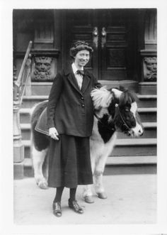 an old black and white photo of a woman standing next to a small cow in front of a building