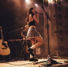 a woman singing into a microphone while standing next to an acoustic guitar on the stage