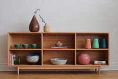 a wooden shelf with bowls, vases and books on it in front of a white wall