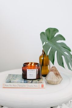 a candle and some books on a white table with a green plant in the background