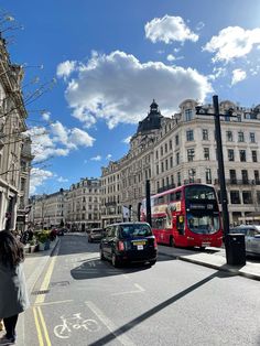 a red double decker bus driving down a street next to tall buildings and parked cars