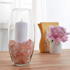 a white candle is sitting in a glass vase with pink rocks on the table next to it