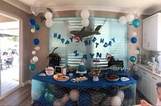 a birthday party with blue and white decorations, balloons and food on a table in front of a window