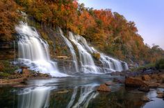 a waterfall with fall foliage surrounding it and the water reflecting its colors in the foreground