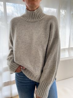 a woman standing in front of a window wearing a gray sweater and jeans with her hands on her hips