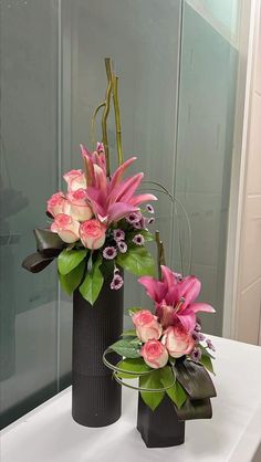 two black vases with pink flowers in them on a white countertop next to a mirror