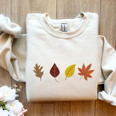 a white shirt with three leaves on it and flowers next to it in front of a wooden floor