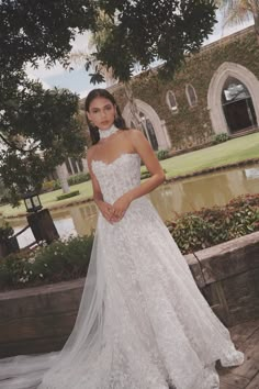 a woman in a white wedding dress standing on a brick wall near a pond and trees