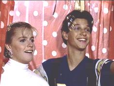 two people standing next to each other in front of a polka dot curtain and holding wine glasses