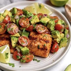 a white plate topped with chicken, avocado and tomato salad next to sliced limes