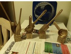 there are some metal utensils on top of a table with newspaper and fan in the background