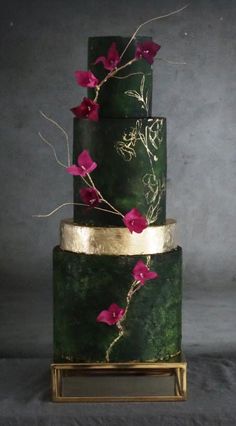 a three tiered green and gold cake with pink flowers on the top, in front of a gray background