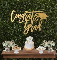 a table topped with a cake and cupcakes next to a wall covered in greenery