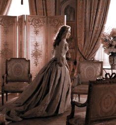 a woman in an elegant dress is standing by a chair and looking out the window