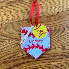 a baseball ornament hanging from a red ribbon on a wooden floor with a name tag