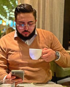 a man sitting at a table with a cup and cell phone in his hand while looking at the screen