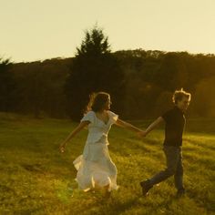 a man and woman holding hands while walking through a grassy field at sundown with trees in the background