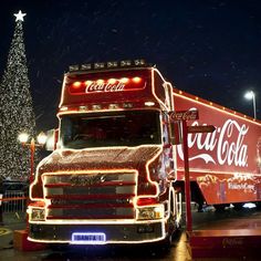 a coca - cola truck parked in front of a christmas tree with lights on it