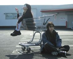 two people sitting on the ground in front of a shopping cart and one person standing up