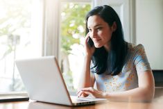 a woman sitting at a table with a laptop computer in front of her, talking on the phone