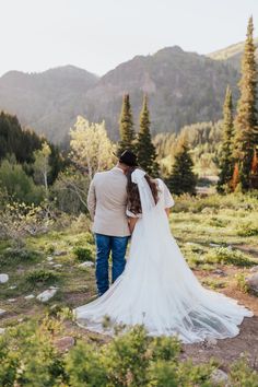 a bride and groom are walking through the woods in their wedding attire, with mountains in the background