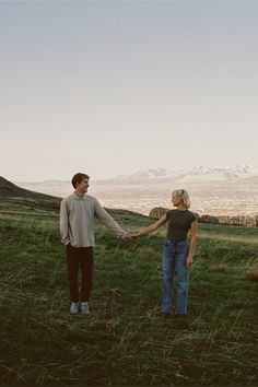 a man and woman holding hands while standing on top of a grass covered hill with mountains in the background