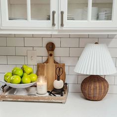 a white kitchen counter topped with green apples and a bowl of fruit next to a lamp