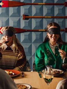 two women wearing blindfolds sitting at a table with food and wine in front of them