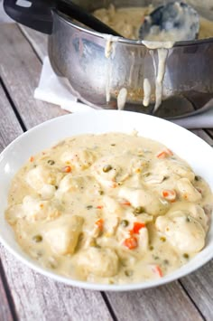 a white bowl filled with chicken and dumplings next to a pot of soup on a wooden table