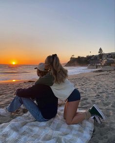 a man and woman sitting on top of a beach next to the ocean at sunset