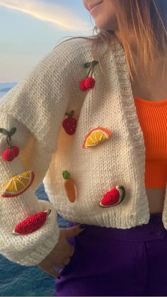 a woman standing next to the ocean with fruit on her sweater