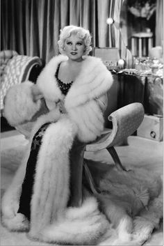 a black and white photo of a woman sitting on a chair wearing a fur coat