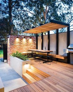 an outdoor dining area with wooden decking and lighting on the outside wall, surrounded by trees