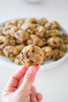 a hand holding up a chocolate chip cookie in front of a plate of other cookies