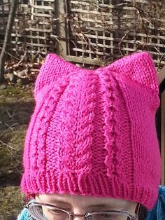 a woman with glasses wearing a pink knitted cat hat and blue sweater in front of a wooden fence