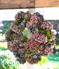 a wreath hanging on the side of a wooden post in front of a window with green leaves and purple flowers