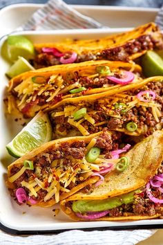 three tacos in a white dish with limes and red onions on the side