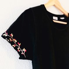 a black t - shirt with pink and yellow flowers embroidered on the chest, hanging from a wooden hanger