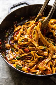 a skillet filled with noodles and vegetables being stirred by tongs on the side