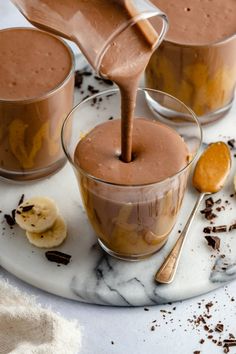 chocolate pudding being poured into two glasses with bananas on the side and spoons next to it