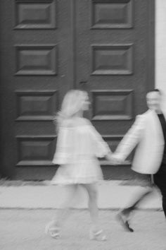 black and white photograph of two people holding hands in front of a building with large doors