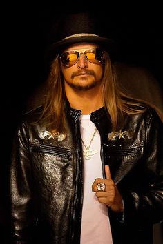 a man with long hair wearing a black leather jacket and hat, giving the peace sign