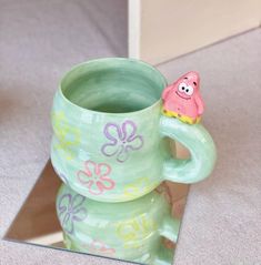 there is a green cup with a pink flower on it and a small figure in the middle