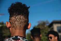 the back of a man's head with tattoos on his face and neck, while two other men stand in the background